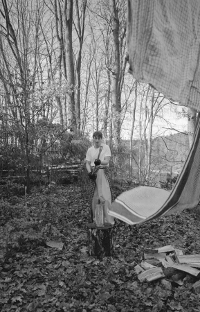 Monochrome outdoor portrait of a young adult standing on a tree stump in a forest holding camera equipment.