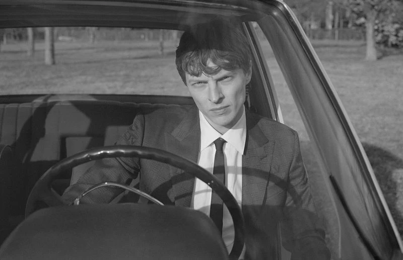 Monochrome portrait of a male sitting in a car, wearing a suit, looking directly at the camera with trees visible in the background.
