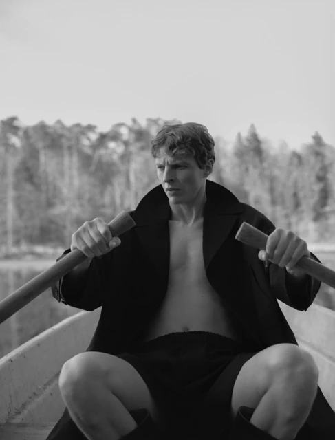 A male rowing a boat on a lake, wearing a black coat and shorts. Forest visible in the background on a bright, overcast day.