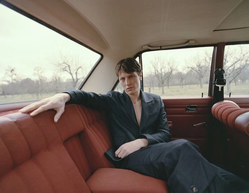 Man in a dark suit sits in the backseat of a vintage car with red leather seats and a view of trees through the back window.