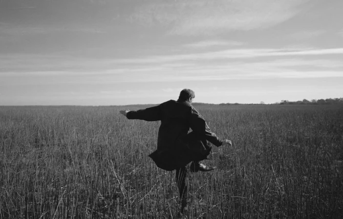 Black and white photo of an adult dancing in a field of tall grass under a cloudy sky, wearing a dark coat and dark shoes.