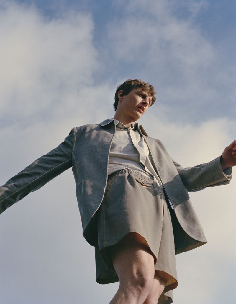 Young man in oversized grey suit and shorts posing against blue sky with white clouds. Fashion shot with natural light.