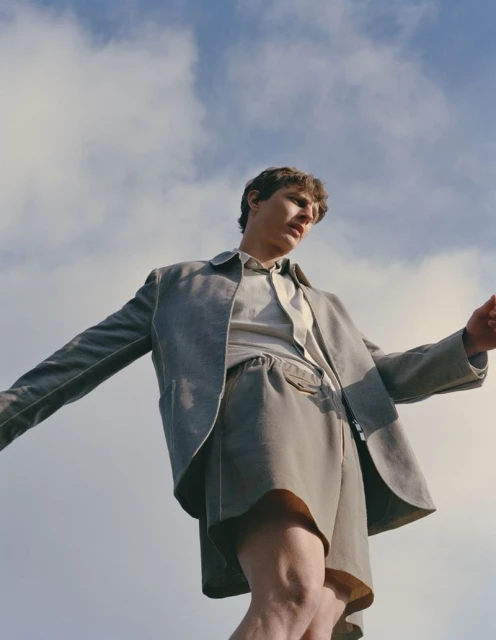 Young man in oversized grey suit and shorts posing against blue sky with white clouds. Fashion shot with natural light.