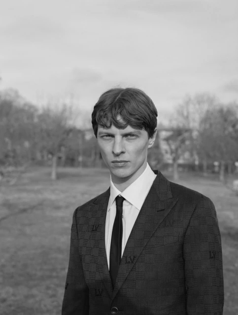 Monochrome portrait of a man in a dark patterned suit with a black tie, standing outdoors in front of a grassy field and bare trees.