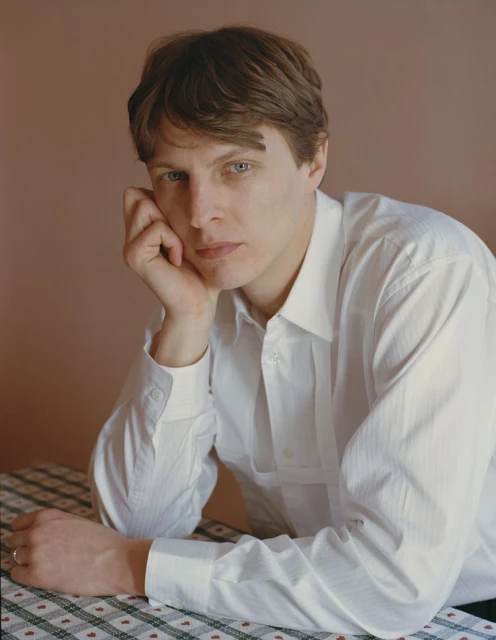 Portrait of an adult male wearing a white button-down shirt leaning on a table with a checkered tablecloth against a peach background.