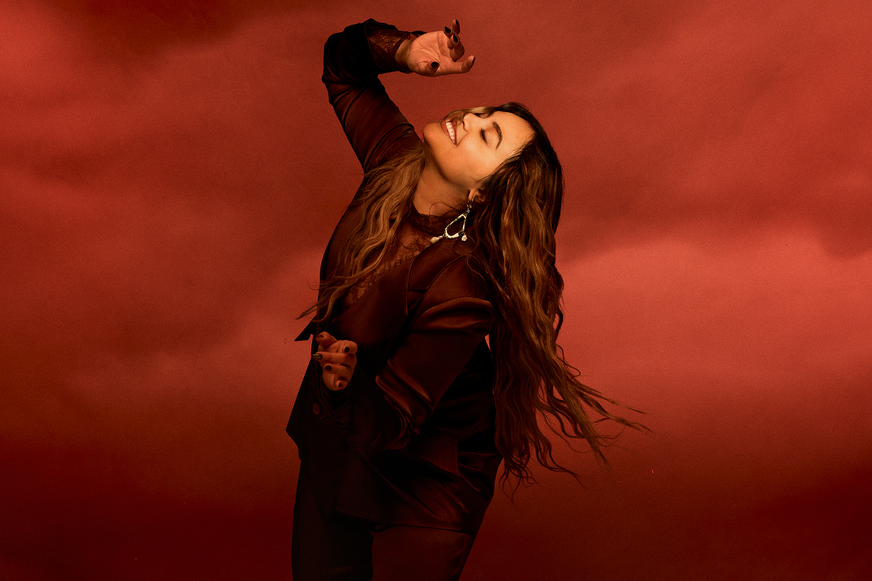 A woman with long brown hair poses in a red monochromatic setting wearing a burgundy suit and statement earrings and smiling.