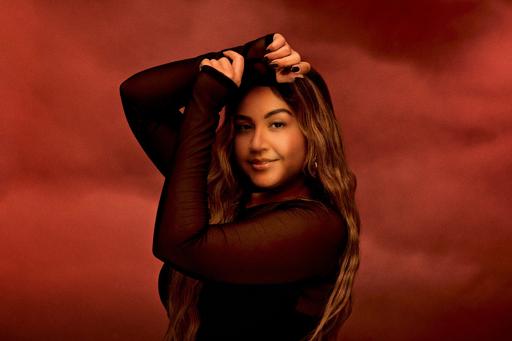 Portrait of a smiling adult woman with long wavy brown hair, wearing a black top, posing against a red studio background with cloud effects.