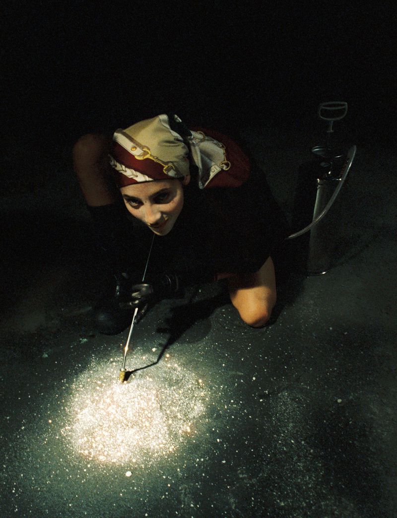 High-angle shot of a woman in a headscarf crouching and holding a tool near glowing powder on a dark surface in an indoor setting.