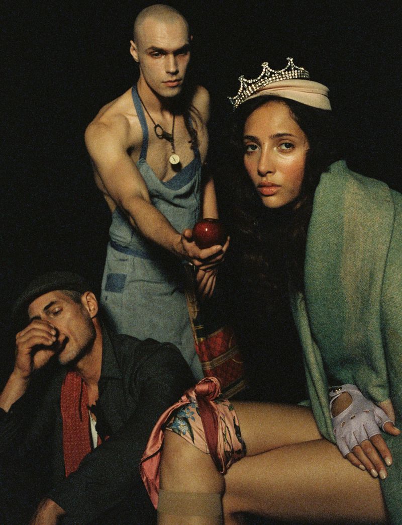 Dramatic fashion portrait features three people in costume: a woman with a tiara, and two men, one holding an apple, against a dark backdrop.