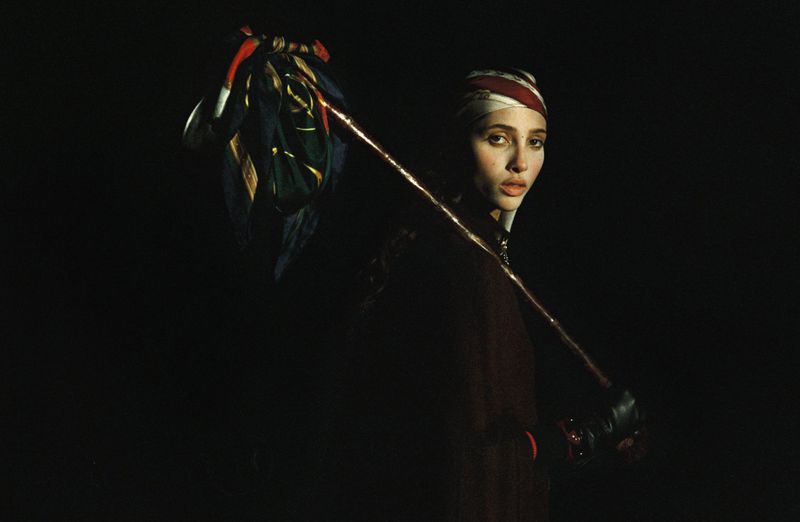 Portrait of a woman in dark clothing and a patterned headscarf, holding a patterned cloth draped over a wooden stick.