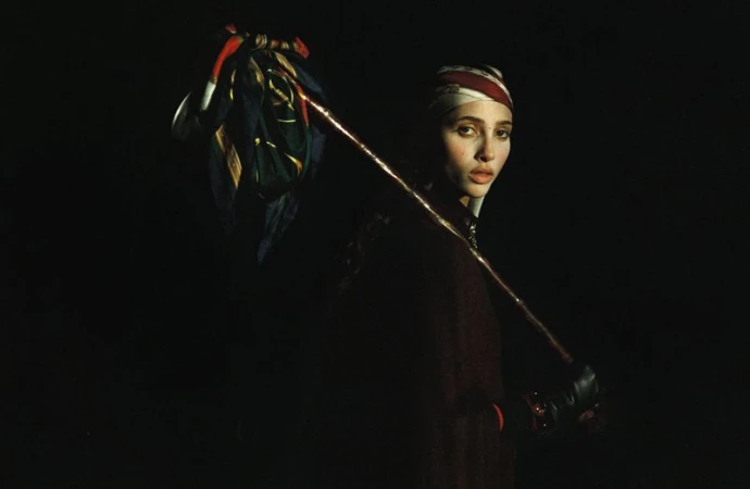 Portrait of a woman in dark clothing and a patterned headscarf, holding a patterned cloth draped over a wooden stick.