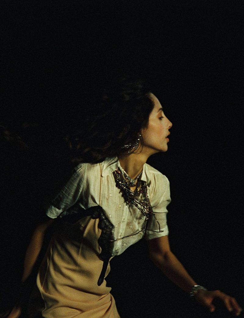 Profile shot of a woman with dark hair wearing a white shirt and layered necklaces with a dark background and soft lighting.