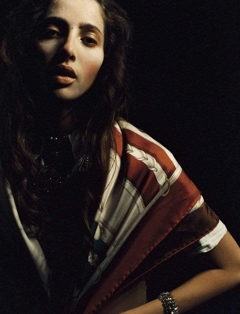 Portrait of a woman with long brown hair, wearing a patterned scarf and jewellery, captured in a dimly lit, high-contrast studio setting.