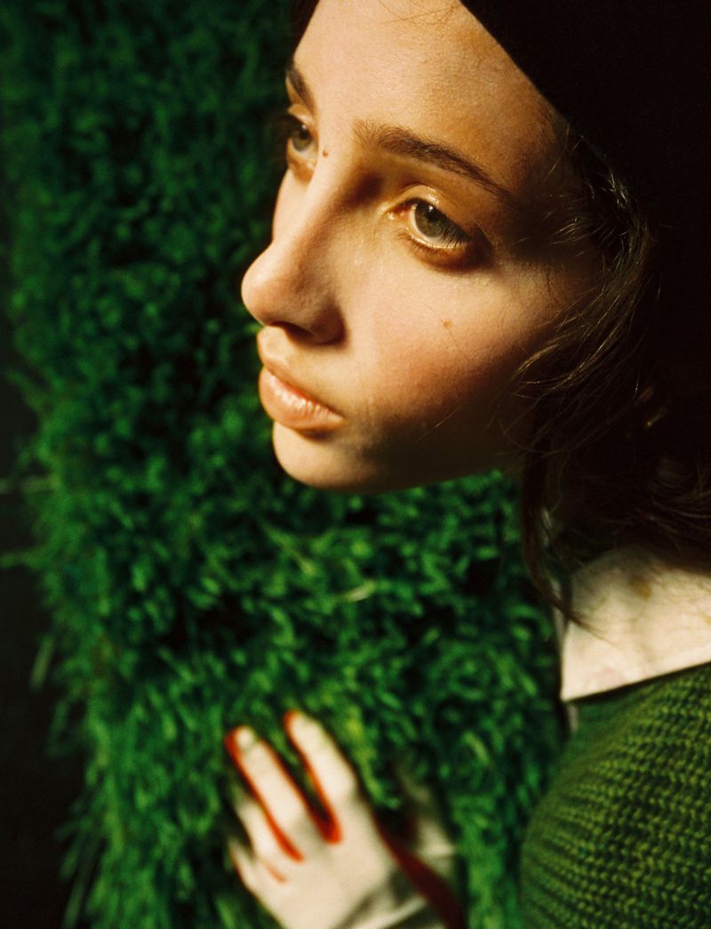 Profile view of a young woman with green eyes. She wears a green sweater and black cap. Her hand, covered in red paint, rests on foliage.