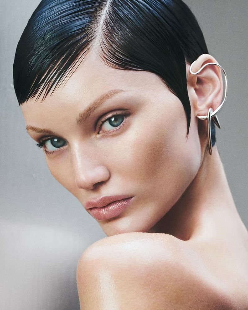 Close-up portrait of a woman with slicked-back dark hair and dramatic silver ear cuff, gazing directly at the viewer in a beauty shot.