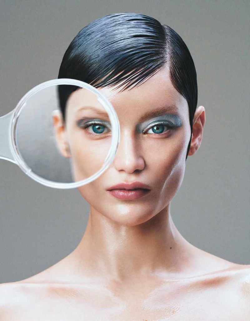 Woman with slicked-back hair and blue eyeshadow poses with a magnifying glass in a beauty shot on a grey backdrop.