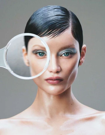 Woman with slicked-back hair and blue eyeshadow poses with a magnifying glass in a beauty shot on a grey backdrop.
