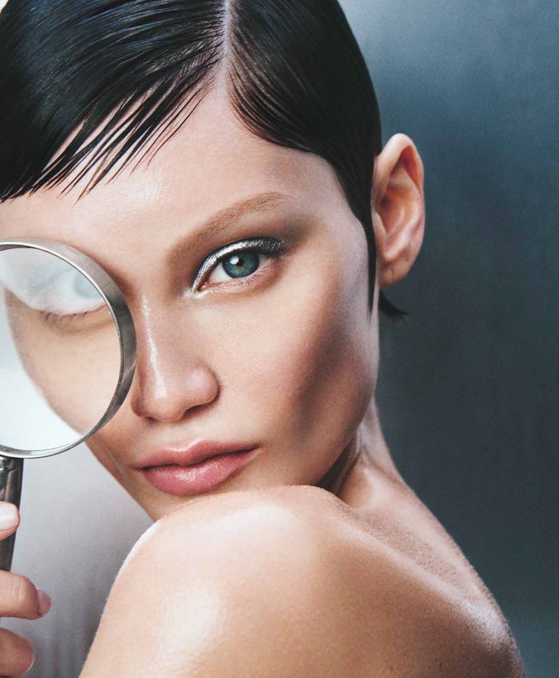 Close-up portrait of a woman with slicked-back hair and silver eyeshadow holding a magnifying glass over her face against a gray background.