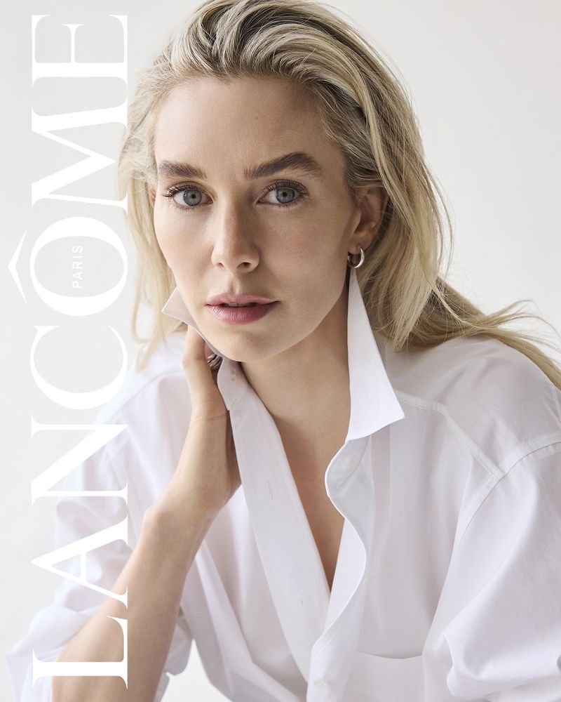 Close-up of a blonde woman in a white shirt. She is fair-skinned, and wears subtle makeup, posing in front of a plain, light background.