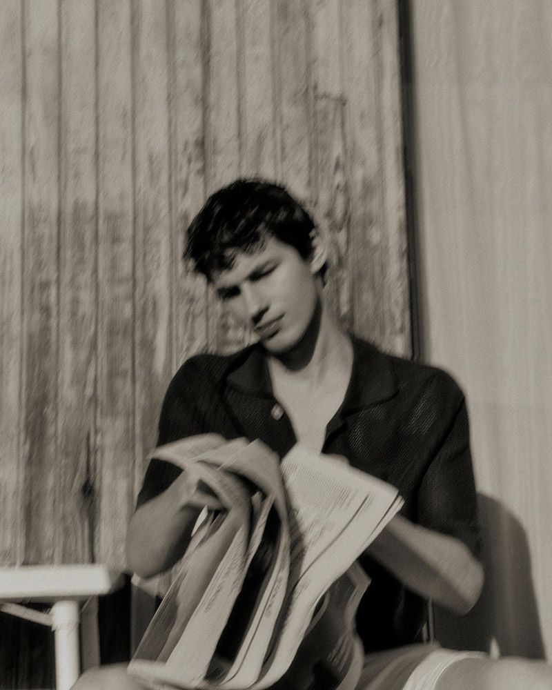 Monochrome portrait of a man reading newspaper outdoors against a wood fence, with short hair, button-down shirt, and soft lighting.