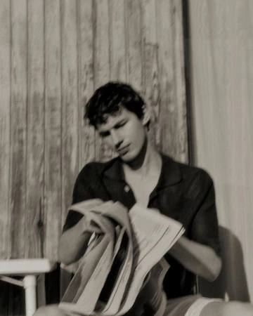 Monochrome portrait of a man reading newspaper outdoors against a wood fence, with short hair, button-down shirt, and soft lighting.
