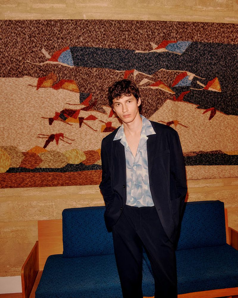 Portrait of a young adult male in a suit and floral shirt, posing in front of an abstract tapestry and blue couch in an indoor setting.