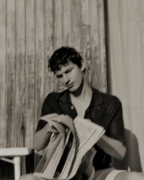 Man wearing dark short sleeve shirt holding open newspaper against a textured wooden slat wall background