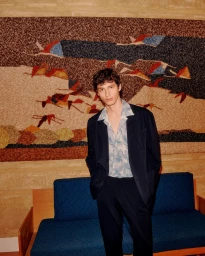 A male model wearing a dark blazer over a light patterned shirt standing in front of a textured tapestry and blue sofa.