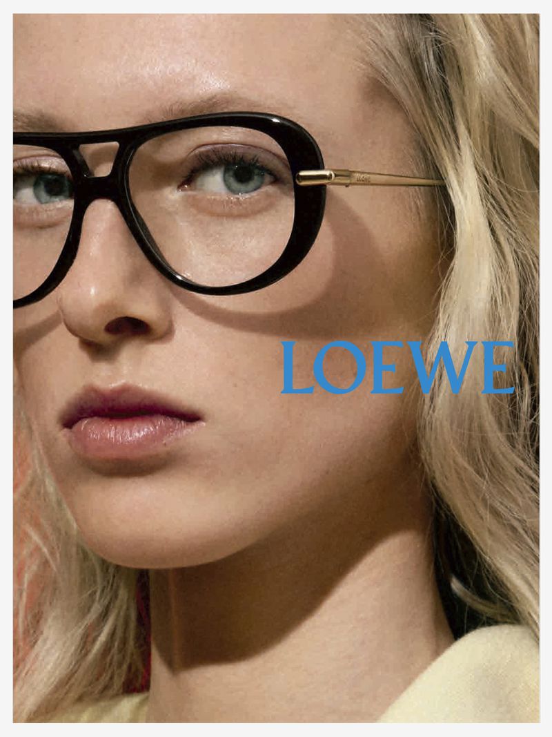 Close-up fashion shot of an adult female model wearing black aviator glasses, light blond hair, and Loewe branding in turquoise. 