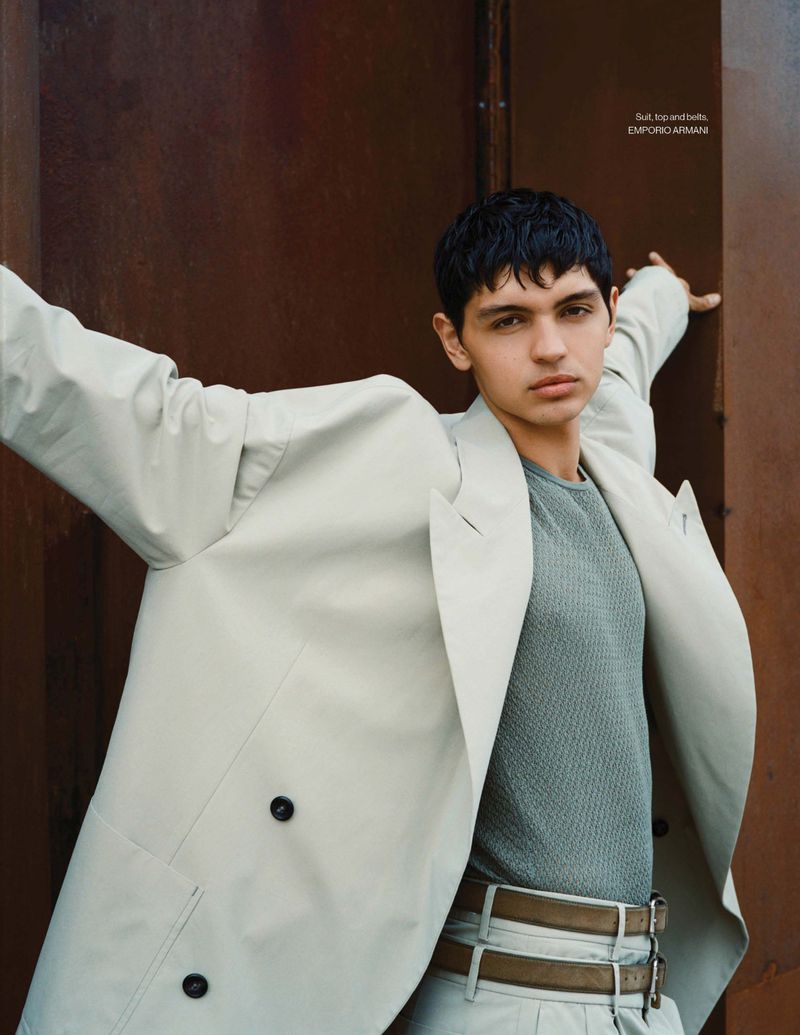 A brunette man poses outdoors wearing a light tan trench coat over a light green shirt and tan pleated pants with a double belt.