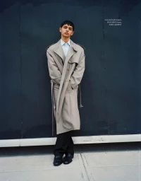 Model wearing an oversized beige trench coat over a light shirt and dark trousers leaning against a dark wall