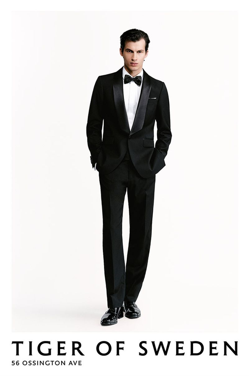 A man in a black tuxedo with a bow tie stands against a white background in a full shot showcasing formal fashion attire.
