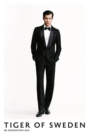 A man in a black tuxedo with a bow tie stands against a white background in a full shot showcasing formal fashion attire.