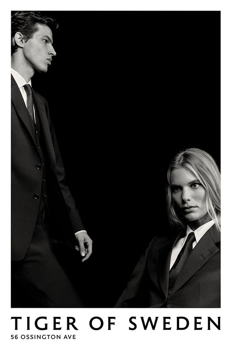 Fashion advertisement featuring a male and female model wearing suits against a dark backdrop, showcasing modern tailoring and design.