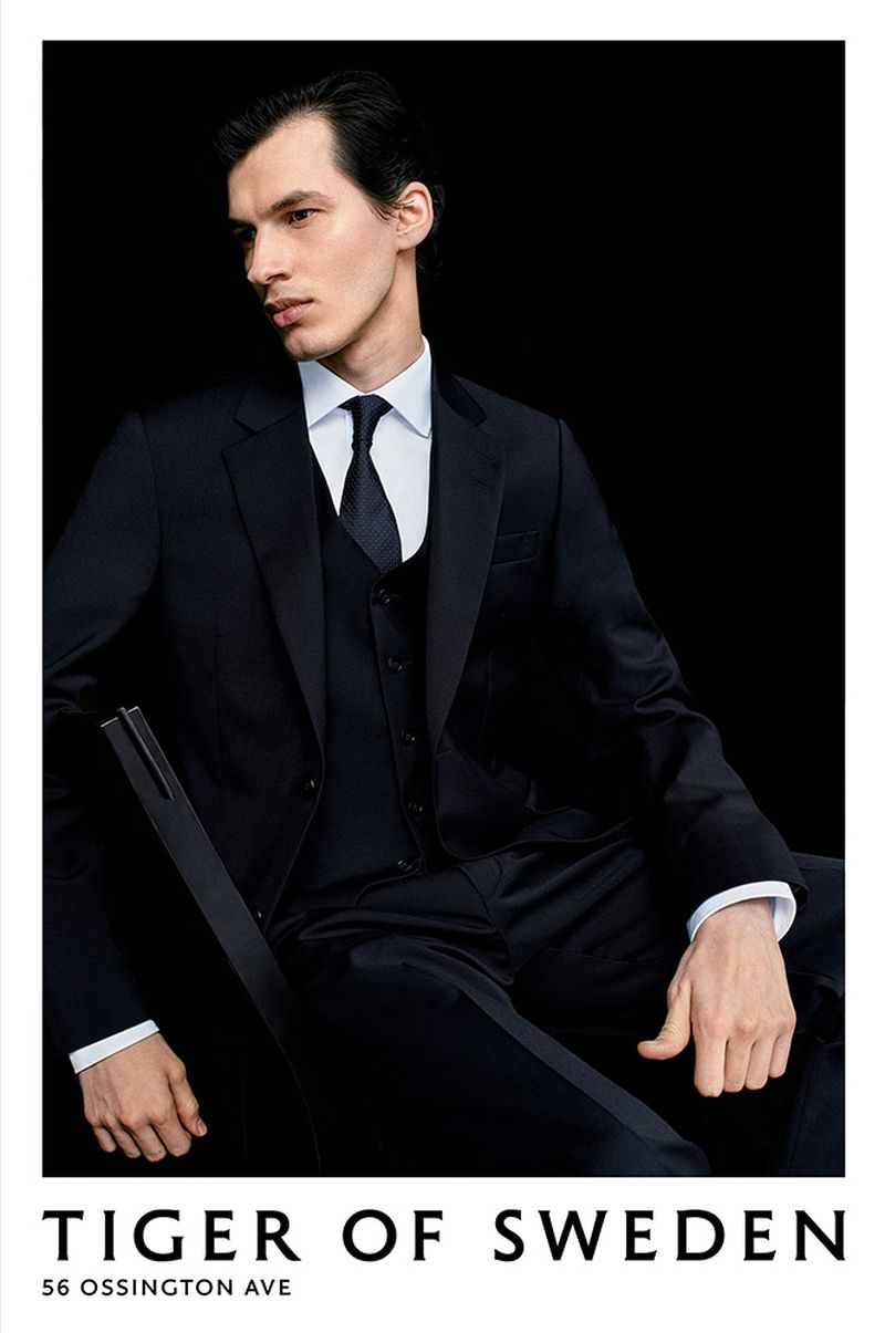 Man in a dark suit with tie and white shirt in a studio shot. The model is seated in a relaxed pose, highlighted against a black backdrop.