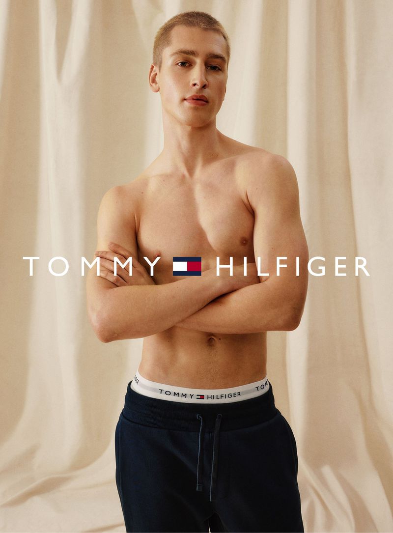 Topless man in navy sweatpants stands in front of beige backdrop in a medium shot, showing branded underwear and Tommy Hilfiger text.
