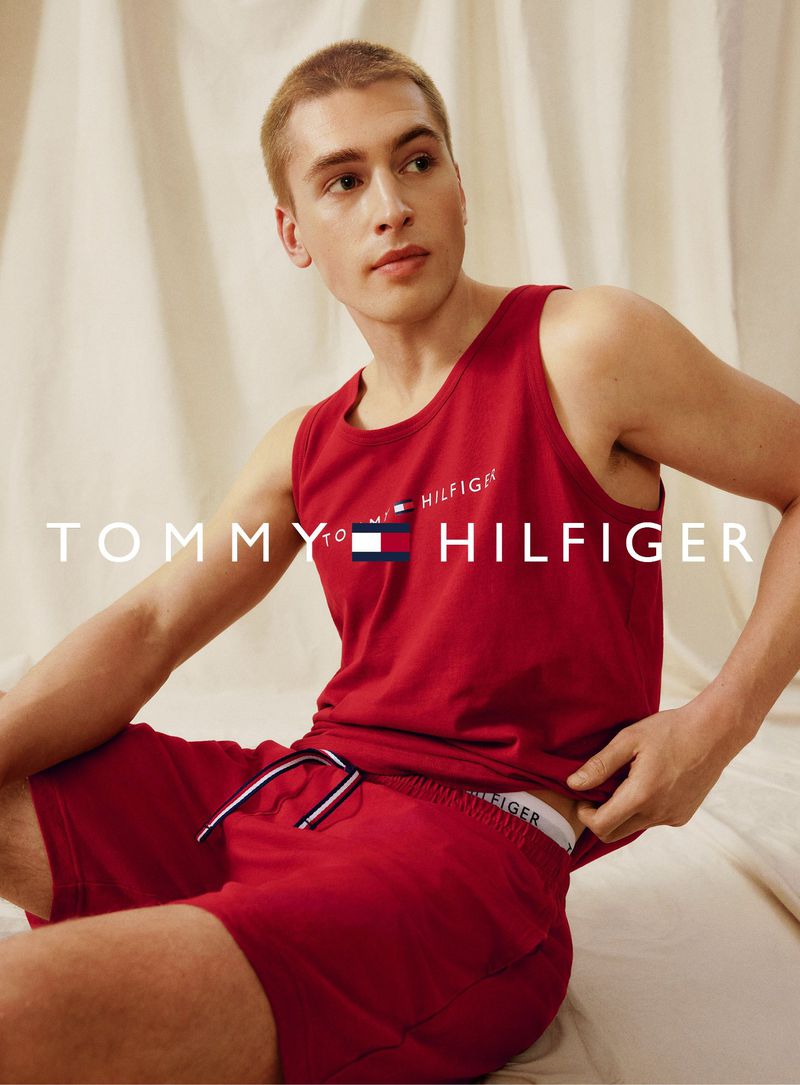 A young adult male sits indoors wearing a red Tommy Hilfiger tank top and shorts, in a studio shot with a soft, neutral background.