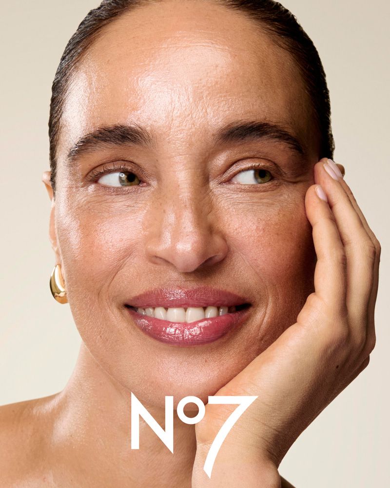 Close-up beauty portrait of a smiling woman with glossy lips and gold hoop earrings against a neutral background.