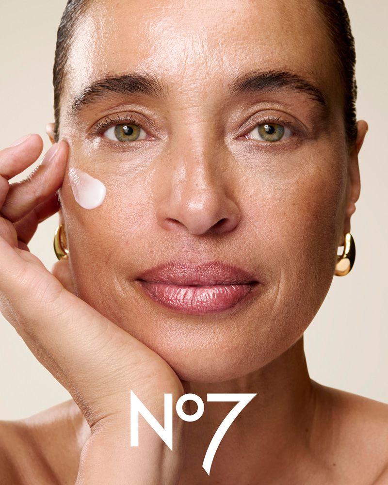 Close-up beauty shot of a woman with light skin, cosmetic cream on her face, and wearing gold earrings. The No7 logo is in the foreground.