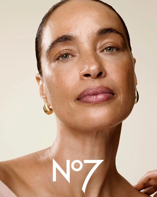 Close-up shot of a woman with gold hoop earrings, wearing makeup with the No7 logo visible on the neck, in a studio setting.