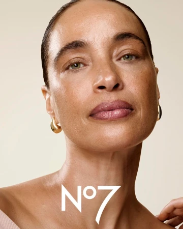 Close-up shot of a woman with gold hoop earrings, wearing makeup with the No7 logo visible on the neck, in a studio setting.