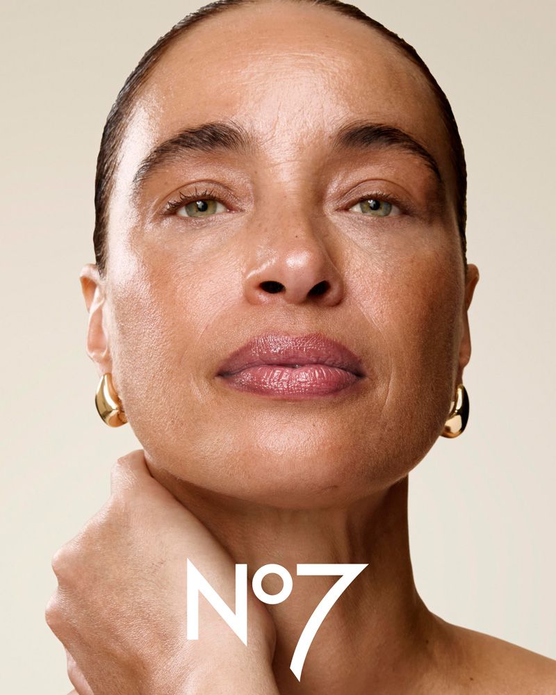 Close-up shot of an adult female model with smooth skin, gold earrings, and the No. 7 logo visible, set against a light beige backdrop.