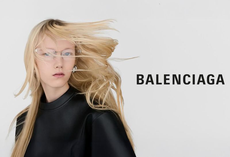Fashion editorial shot of a woman with blonde hair wearing glasses and a black top against a white background with Balenciaga logo.