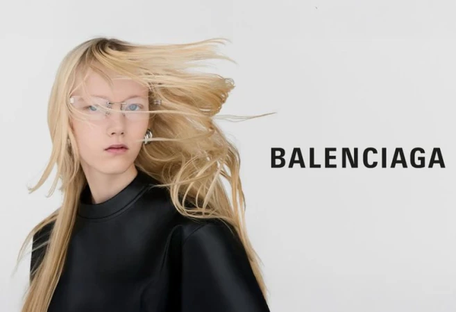 Fashion editorial shot of a woman with blonde hair wearing glasses and a black top against a white background with Balenciaga logo.