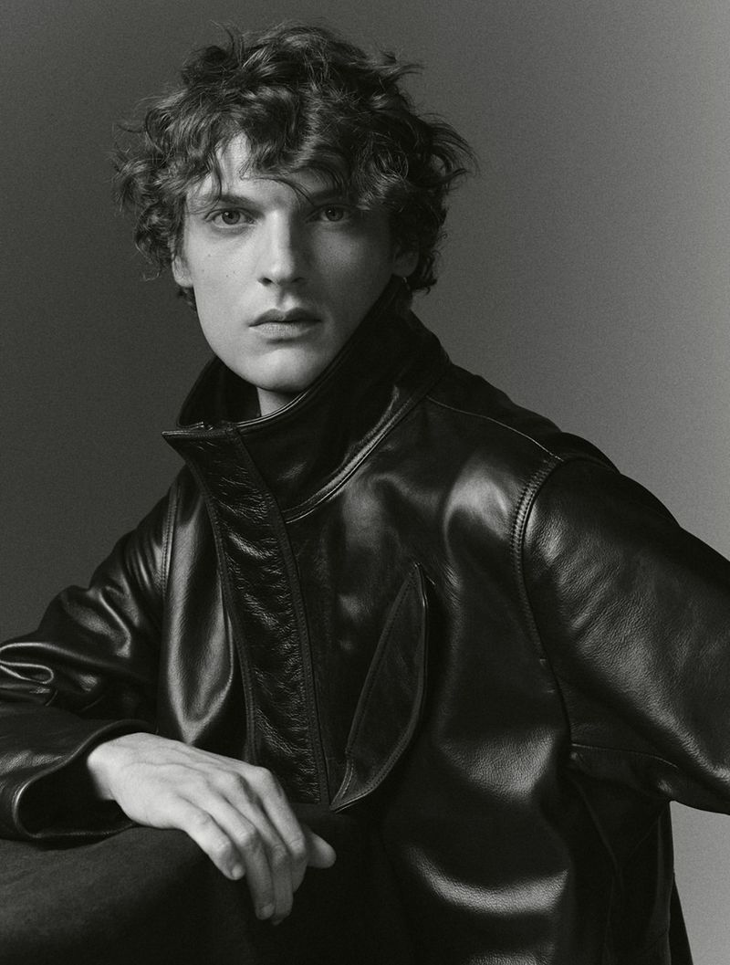 Close-up portrait of a man with curly hair wearing a black leather jacket against a neutral gradient background.