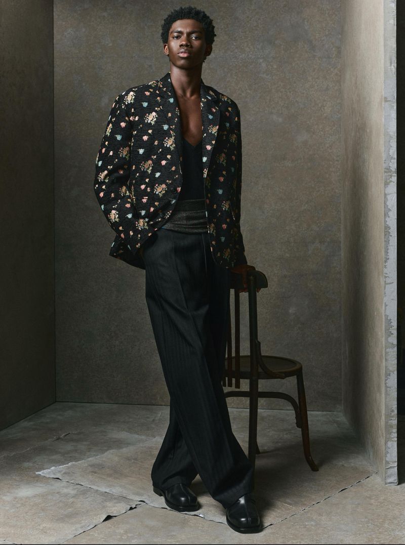 A male model standing leaning against a wooden chair in a dark patterned blazer and wide black trousers.
