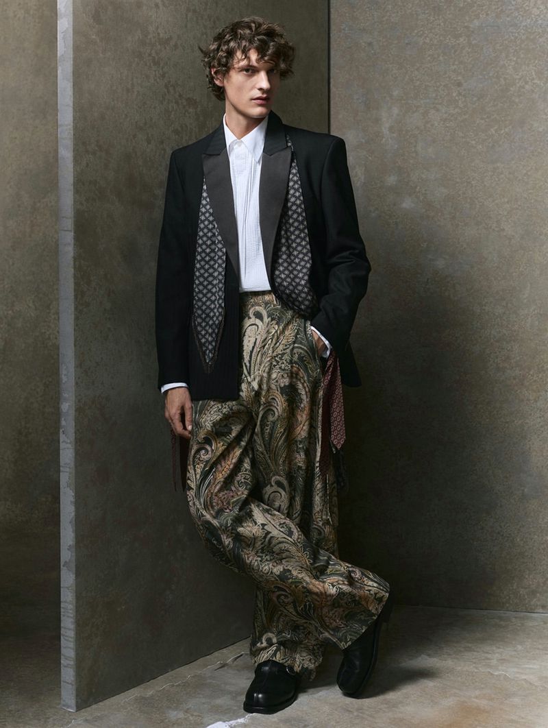 Full-shot fashion portrait of a model in a black blazer, white shirt, and paisley print pants against a neutral-toned studio backdrop.