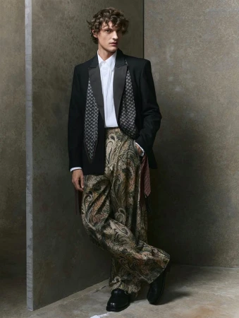 Full-shot fashion portrait of a model in a black blazer, white shirt, and paisley print pants against a neutral-toned studio backdrop.
