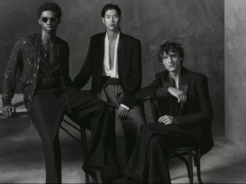 Three fashion models pose in black and white, seated and wearing black suits and open shirts in a high-contrast studio portrait.