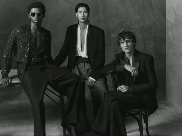Three fashion models pose in black and white, seated and wearing black suits and open shirts in a high-contrast studio portrait.